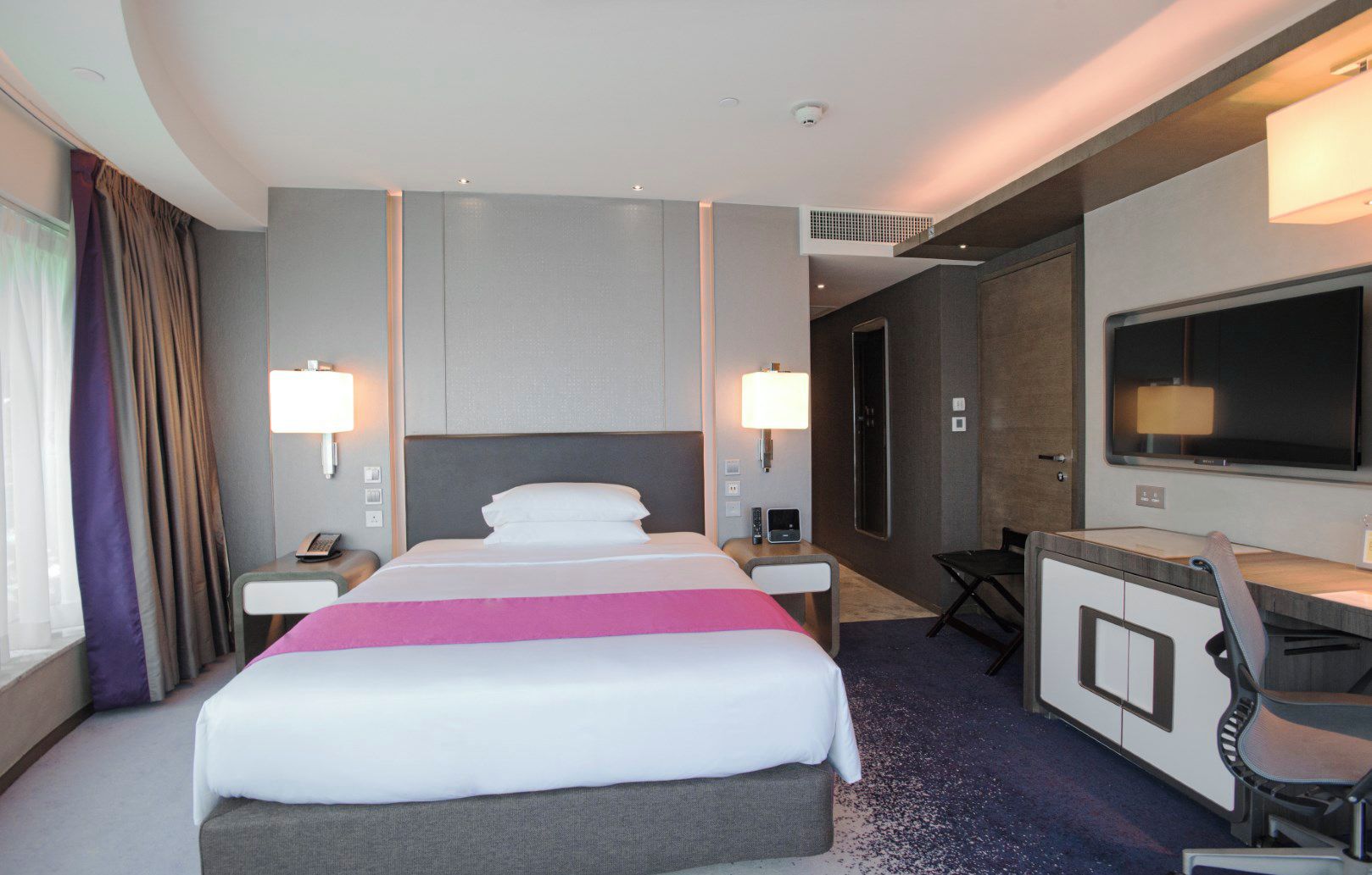 A hotel room with a large bed, bedside tables, a desk with a chair, a TV, and a window with curtains.