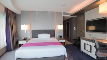 A hotel room with a large bed, bedside tables, a desk with a chair, a TV, and a window with curtains.