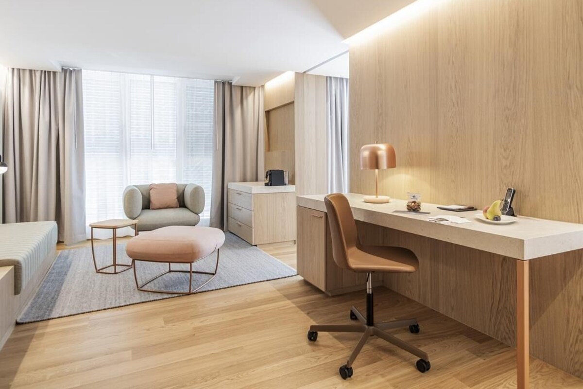 A modern hotel room with a wooden desk, a comfortable sofa, a small side table, and a large window with curtains.