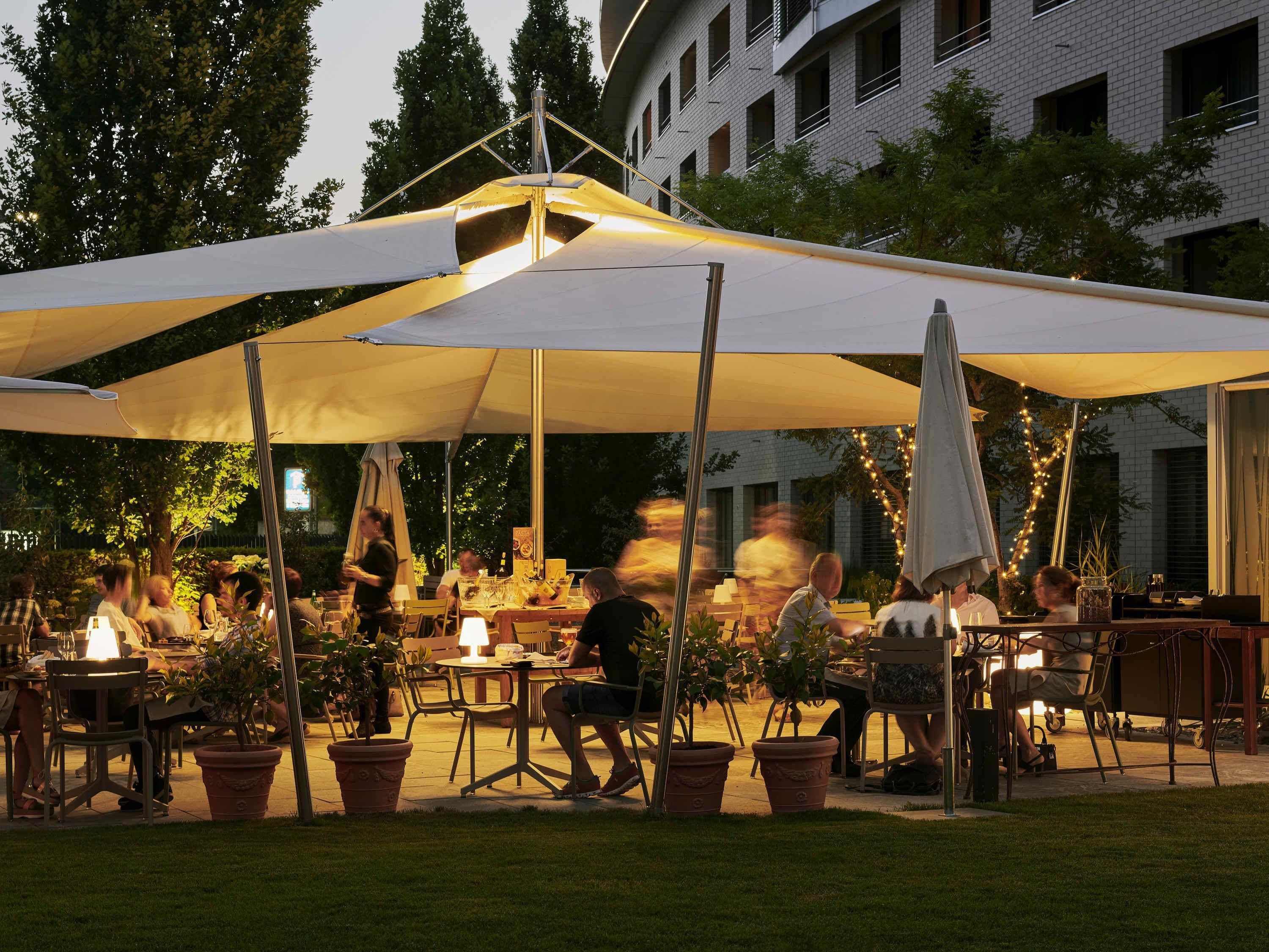 Un espace de restauration en plein air, avec des tables et des chaises, entouré de plantes en pot et doté d’un grand parasol pour offrir de l’ombre.