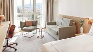 A hotel room with a bed, two sofas, a desk, a chair, a small table, and a view of a cityscape. - Swissôtel Kursaal Bern (Bern)