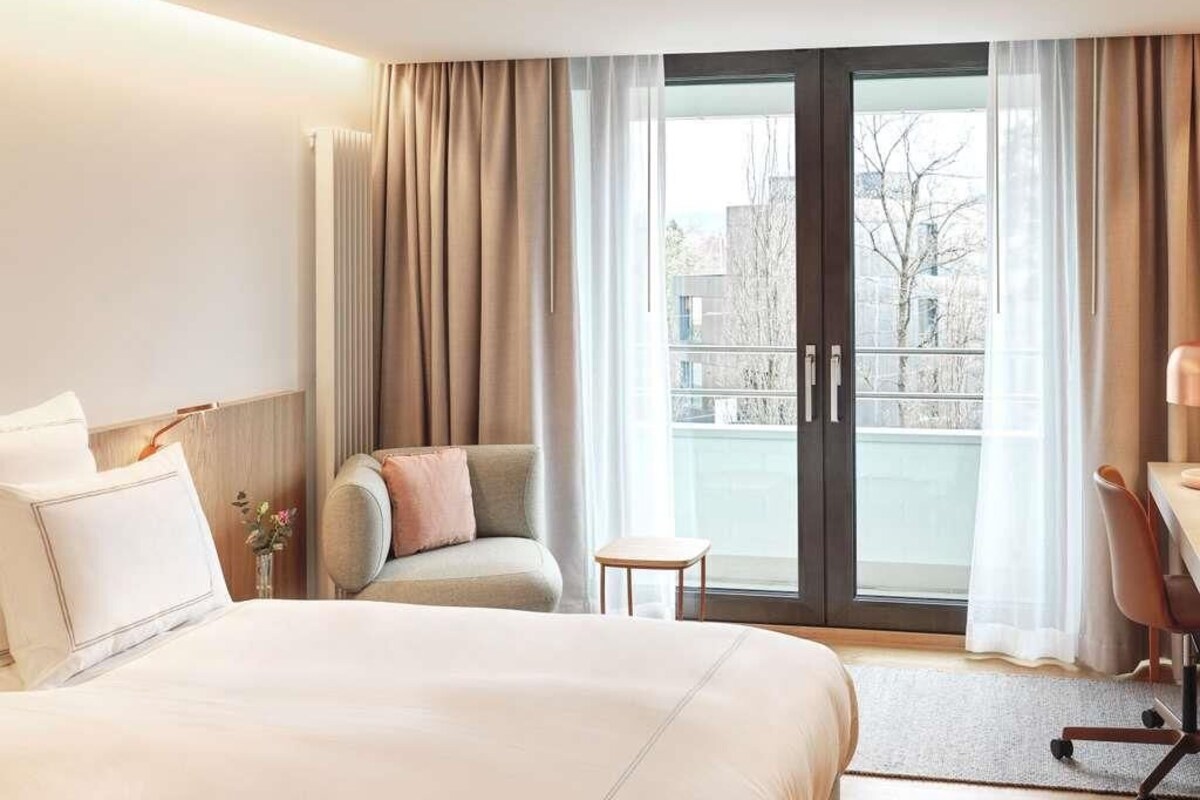 A hotel room with a bed, a chair, a desk, and a view through a glass door.