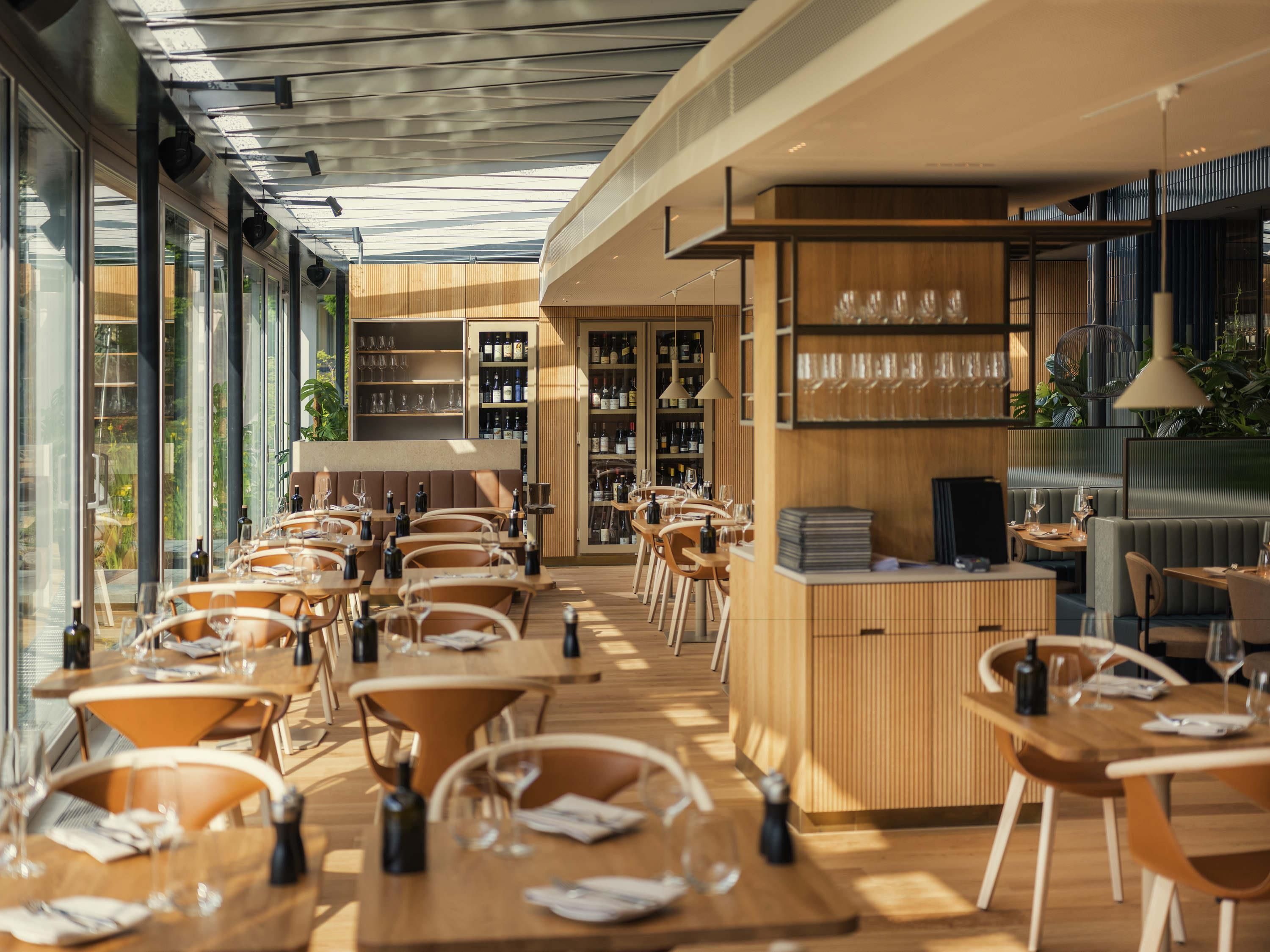 Un interno ristorante moderno con tavoli e sedie in legno, un'area bar con bicchieri da vino e scaffali pieni di bottiglie.