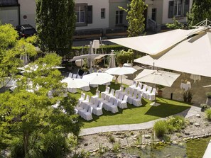 An outdoor seating area with white chairs and umbrellas, surrounded by greenery and a small pond.