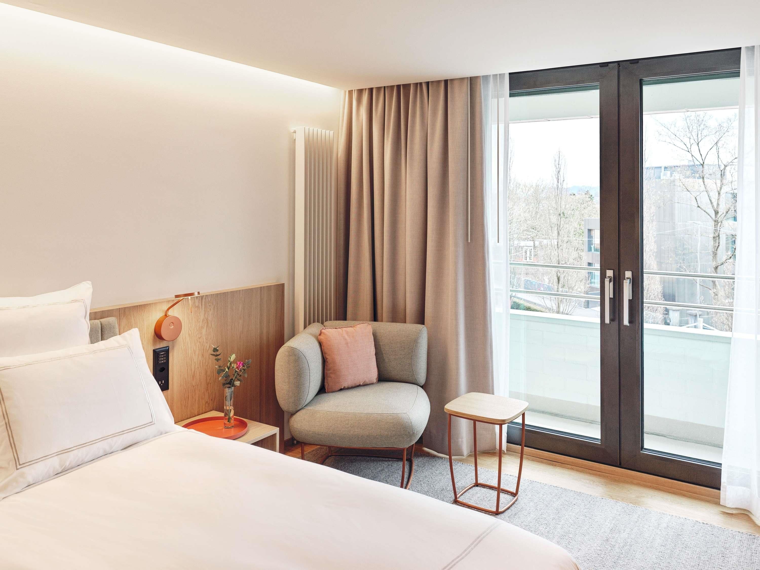A hotel room with a bed, a chair, a small table, a nightstand, and a view through a glass door.