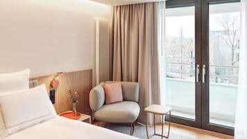 A hotel room with a bed, a chair, a small table, a nightstand, and a view through a glass door.