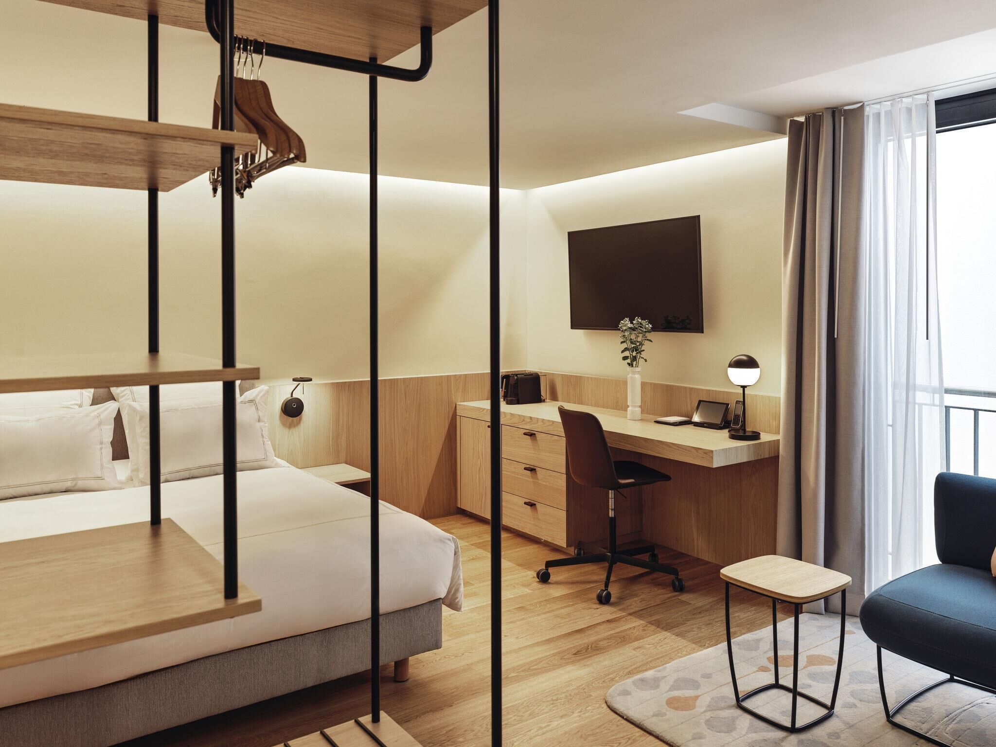 A hotel room with a bunk bed, a desk with a chair, a small table, a TV mounted on the wall, and a window with curtains.