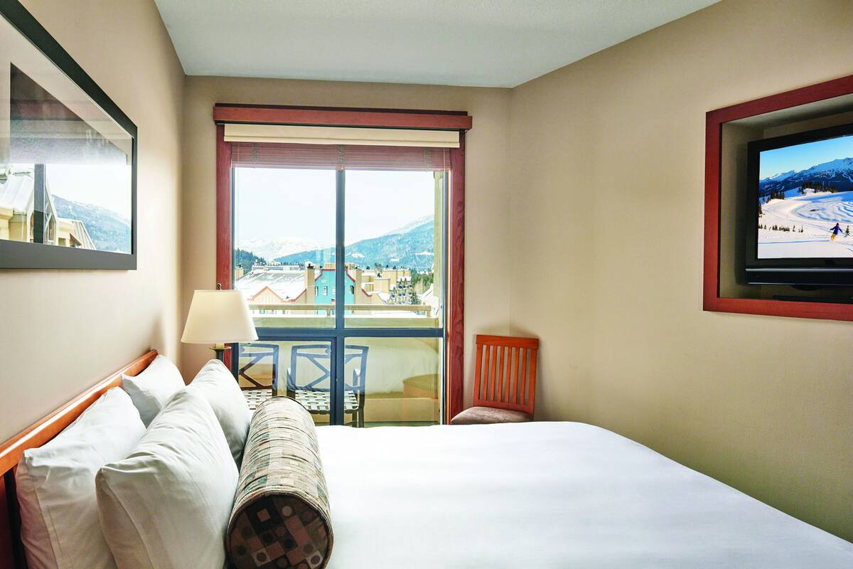Habitación de hotel con una cama grande, un televisor montado en la pared, una ventana con vistas a un paisaje nevado y una silla junto a la ventana.