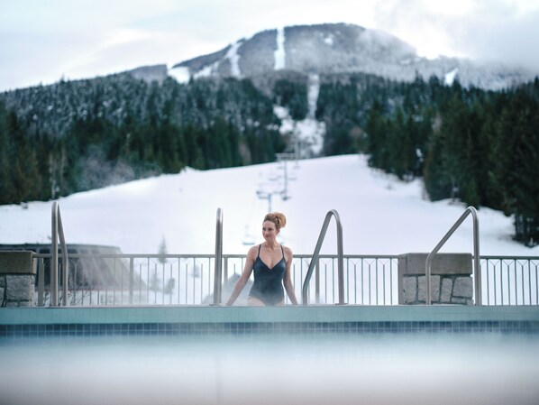 Une personne se trouve au bord d’une piscine, avec une montagne enneigée et un télésiège en arrière-plan.