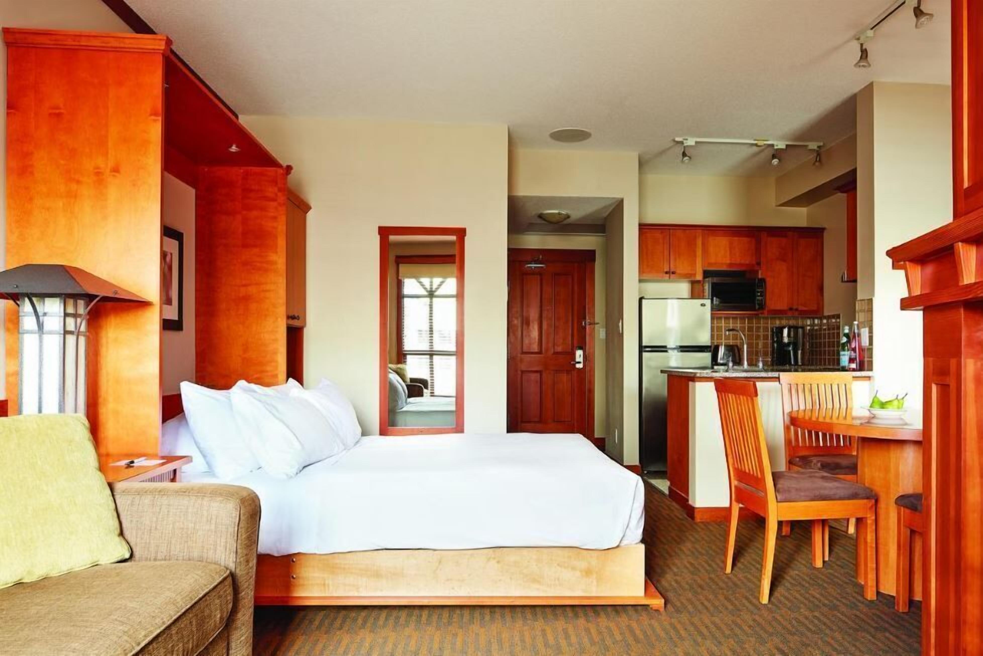 Une chambre d’hôtel comprenant un lit, un canapé, un coin repas avec une table et des chaises, et une cuisine avec des meubles en bois et des appareils électroménagers.
