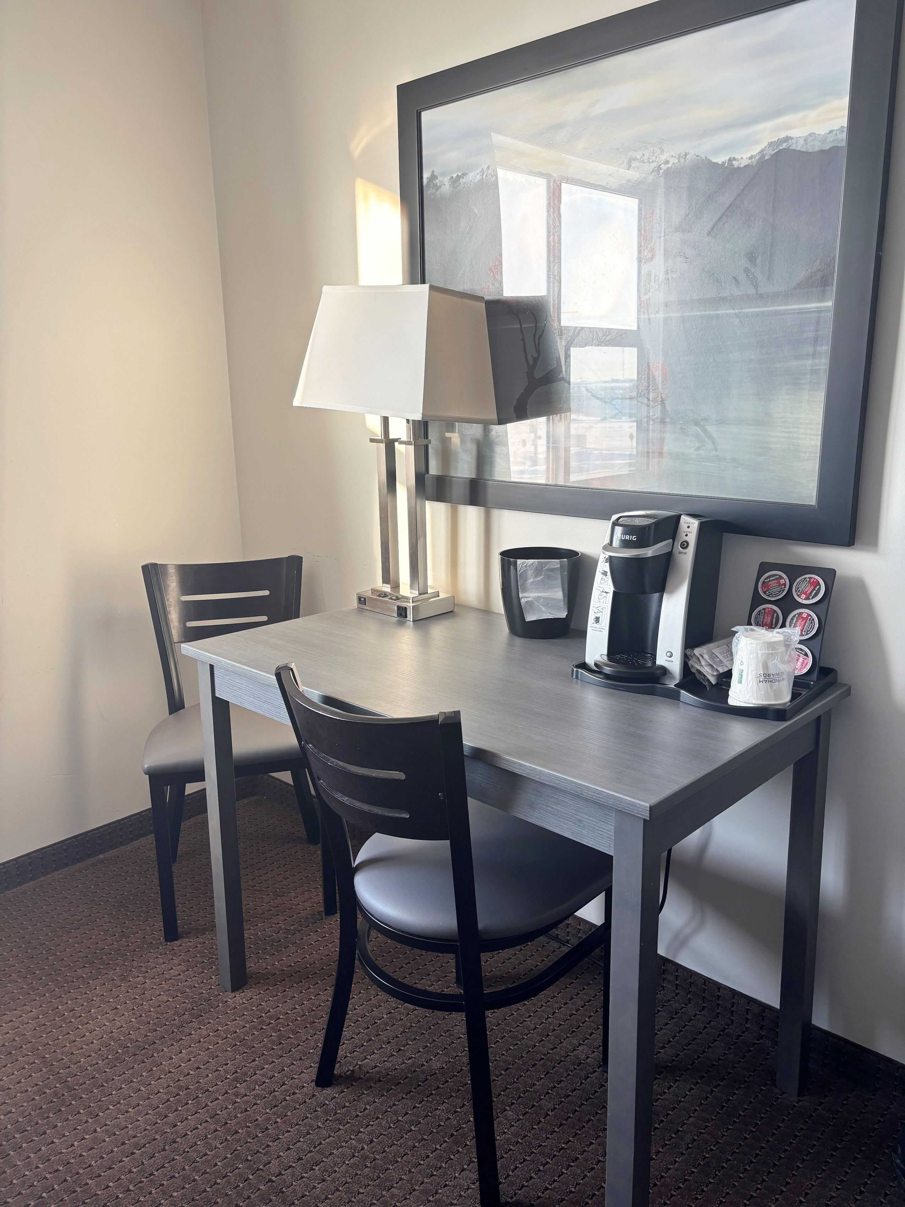 Habitación de hotel con escritorio, una silla, lámpara, cafetera y un cuadro en la pared.