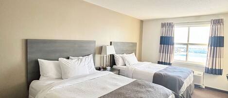 Habitación de hotel con dos camas, una mesita de noche, una lámpara y un ventanal con cortinas.