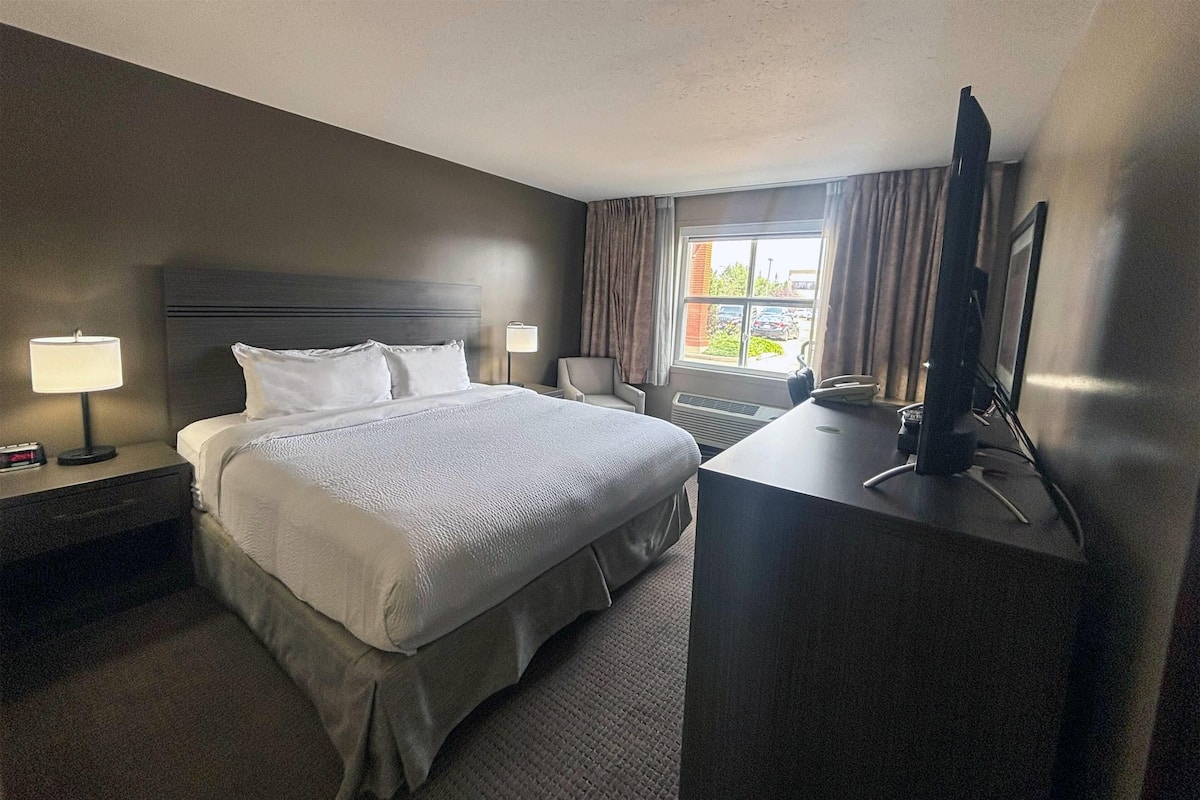 Une chambre d’hôtel avec un grand lit, un bureau avec une télévision, une chaise, une lampe et une fenêtre avec des rideaux.