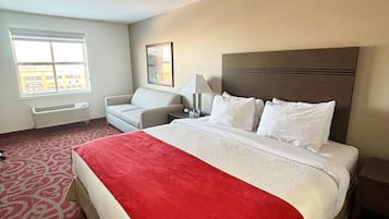 A hotel room with a large bed, a red throw blanket, a white headboard, a bedside table with a lamp, a chair, and a window with a view of buildings.