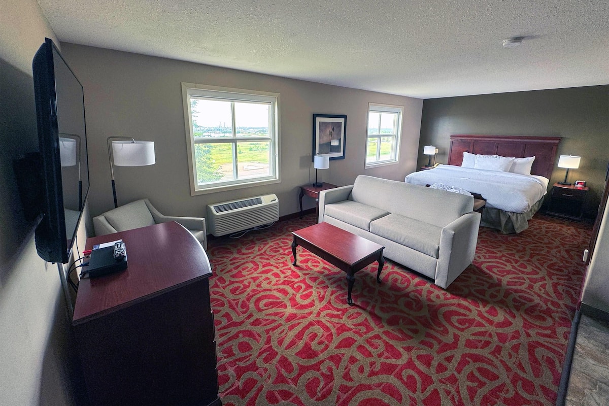 Une chambre d’hôtel avec une moquette à motifs rouges, un lit avec une literie blanche, un canapé, un bureau avec une lampe et une grande fenêtre offrant une vue.