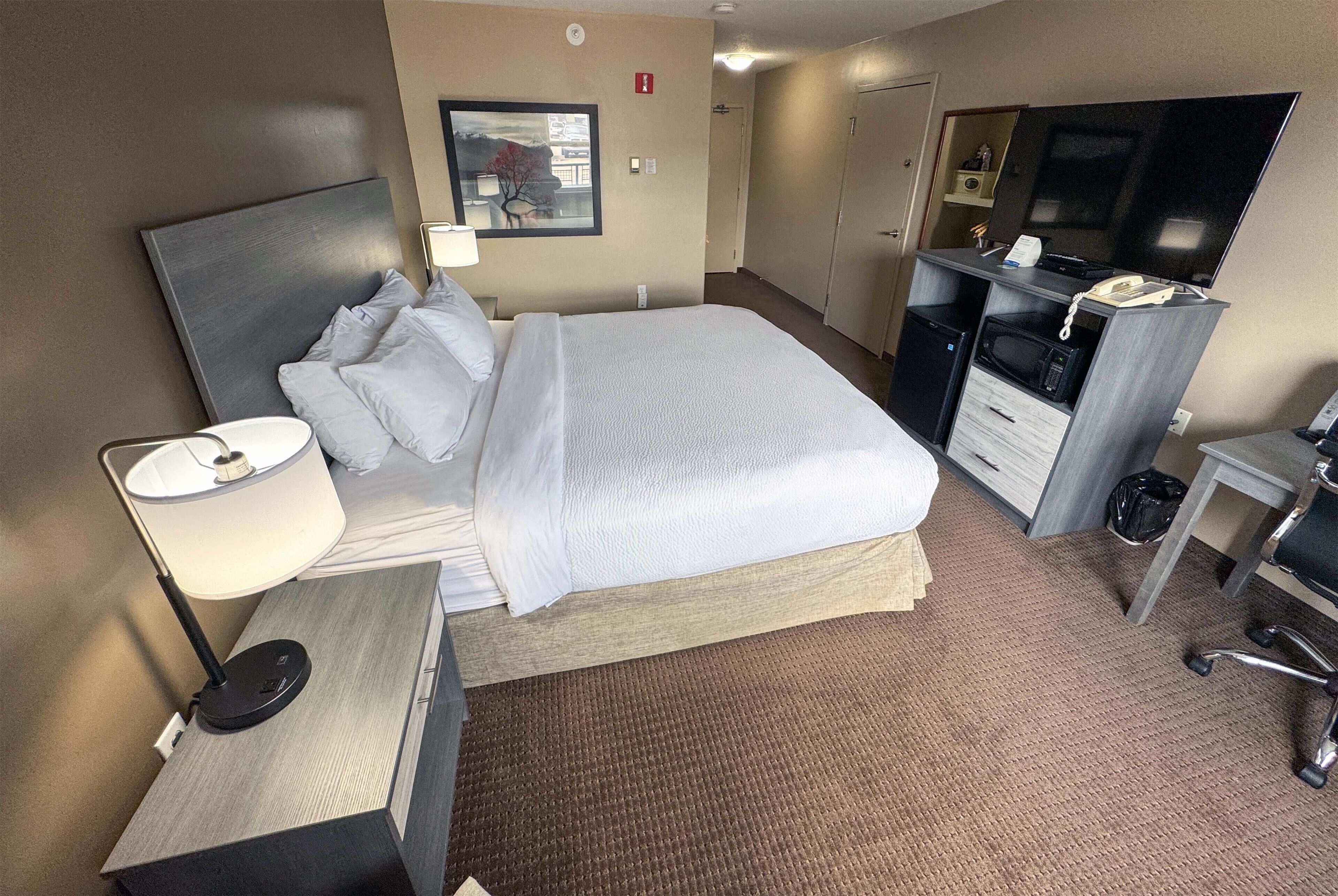 Una habitación de hotel con cama, mesita de noche, lámpara, escritorio, silla, televisor y una pequeña kitchenette.