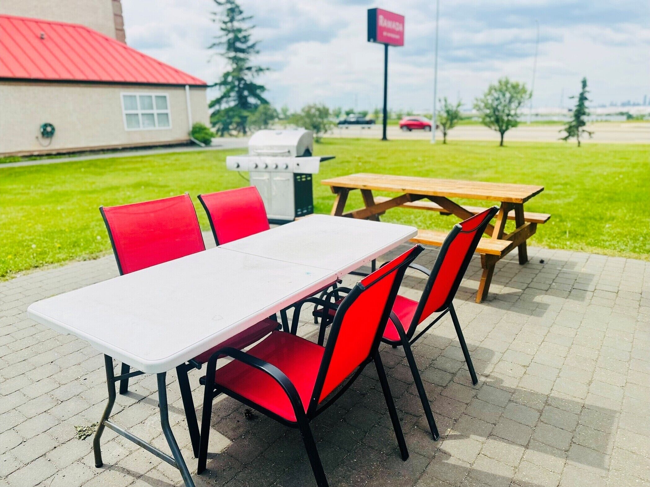 Un área de comedor al aire libre con una mesa blanca y sillas rojas, una parrilla y una mesa de picnic en un entorno similar a un parque.