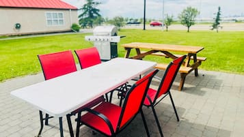 Un área de comedor al aire libre con una mesa blanca y sillas rojas, una parrilla y una mesa de picnic en un entorno similar a un parque.