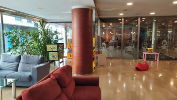 Un vestíbulo moderno con zona de estar, un pilar grande y un escaparate de vidrio con área de juegos infantiles.