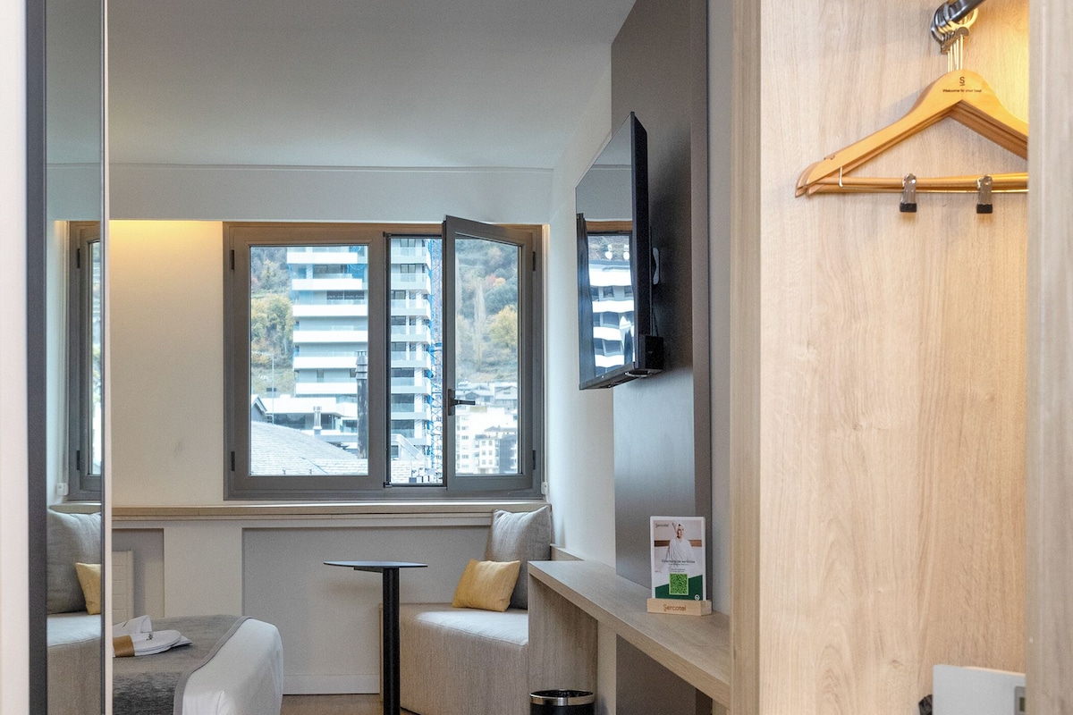 A modern, compact room with a sofa, a small table, a TV mounted on the wall, and a wooden cabinet with a hanging rack.