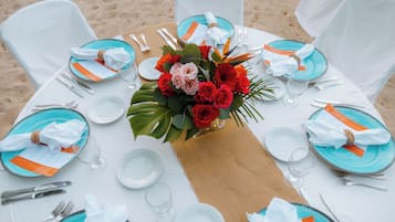 Una mesa redonda preparada para una comida, con platos blancos y azules, cubiertos y un centro de mesa con flores rojas y rosadas.