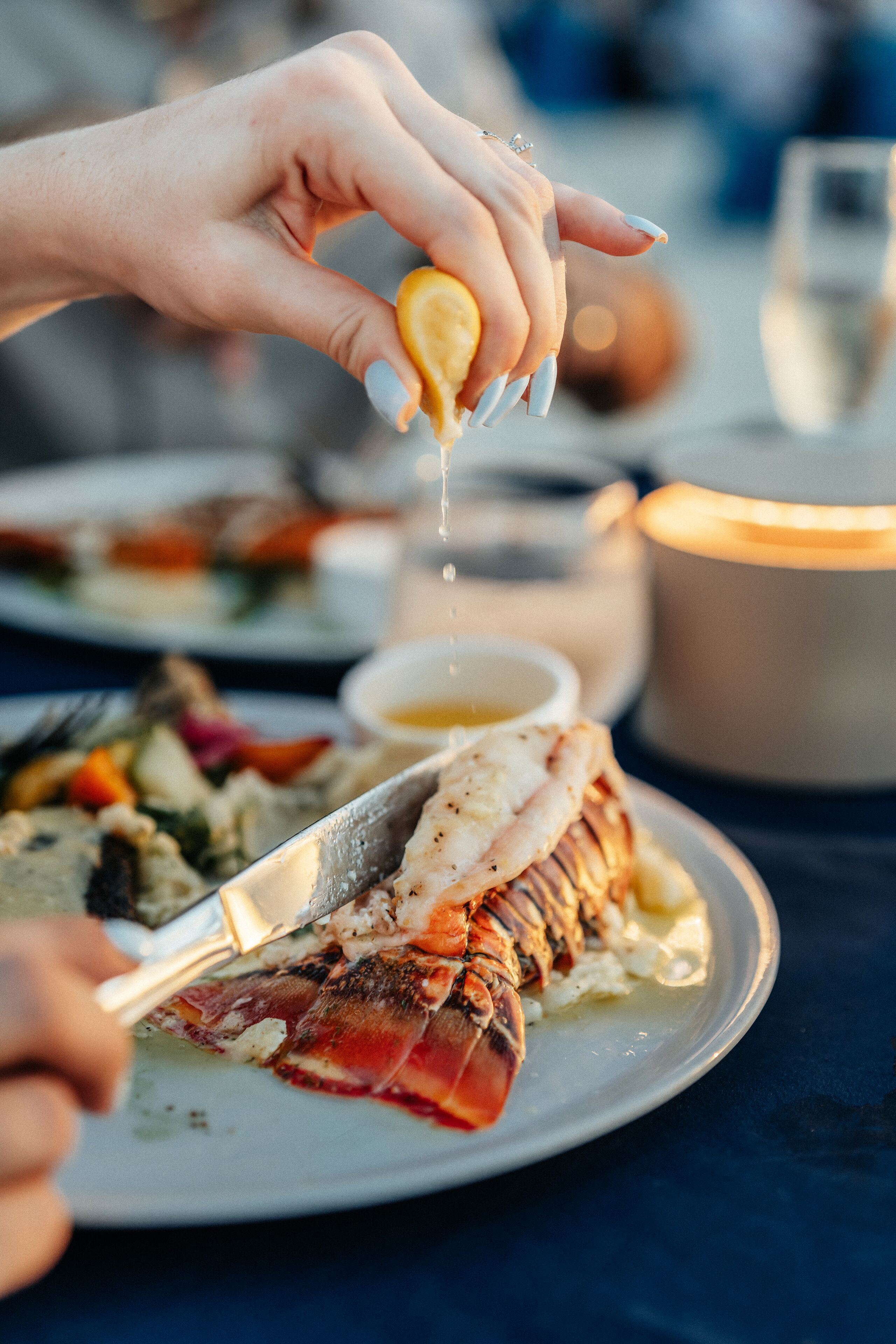 A person holding a lemon over a lobster tail on a plate.