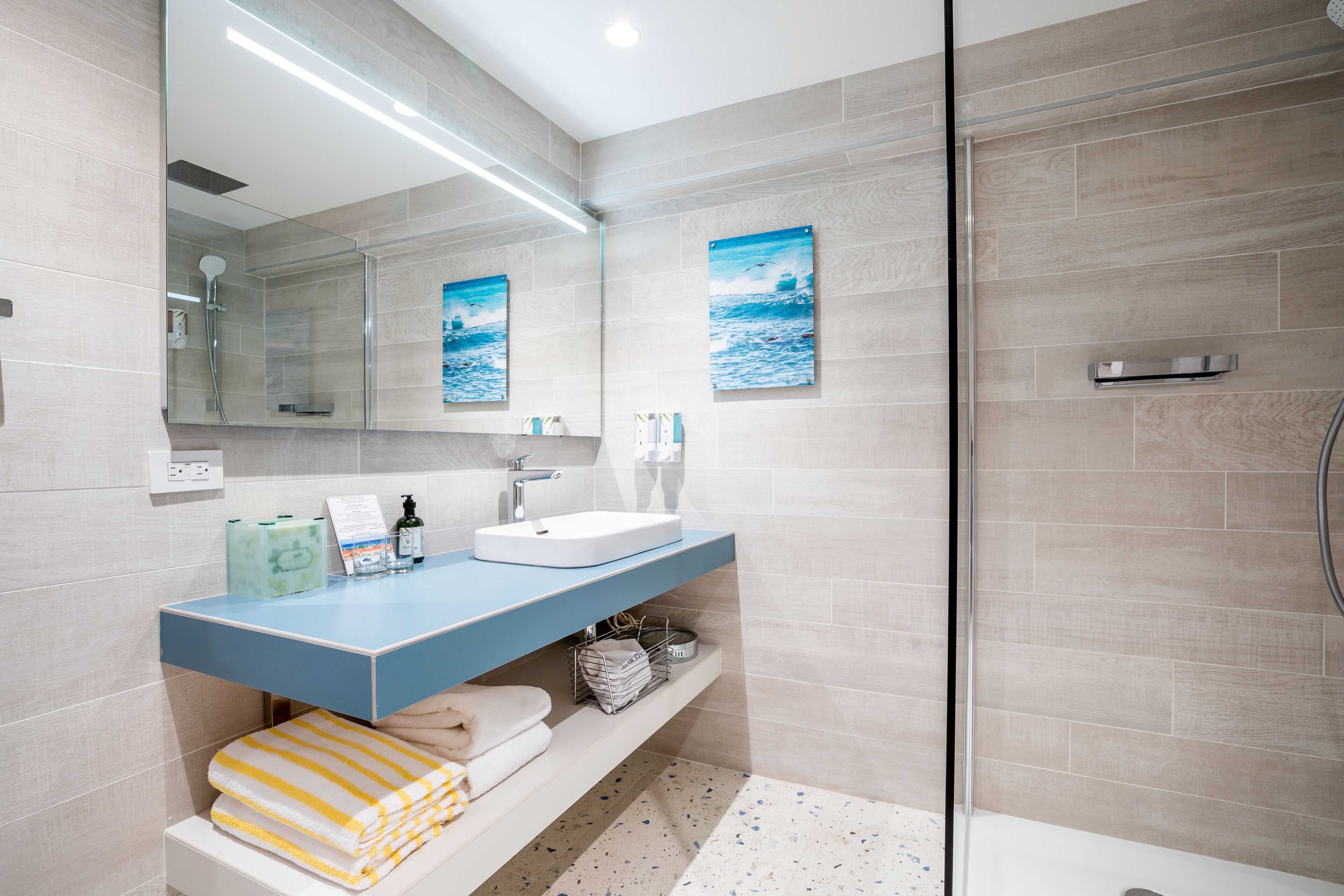 Une salle de bain moderne avec une douche à l’italienne en verre, un plan de toilette bleu et deux tableaux encadrés représentant la mer.