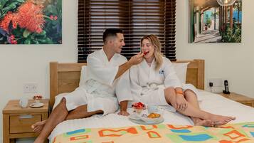 Un couple, vêtu de peignoirs blancs, est allongé sur un lit recouvert d’une courtepointe aux couleurs vives. Sur une table de chevet, on aperçoit une tasse et un bol de fruits. Un tableau orne le mur.