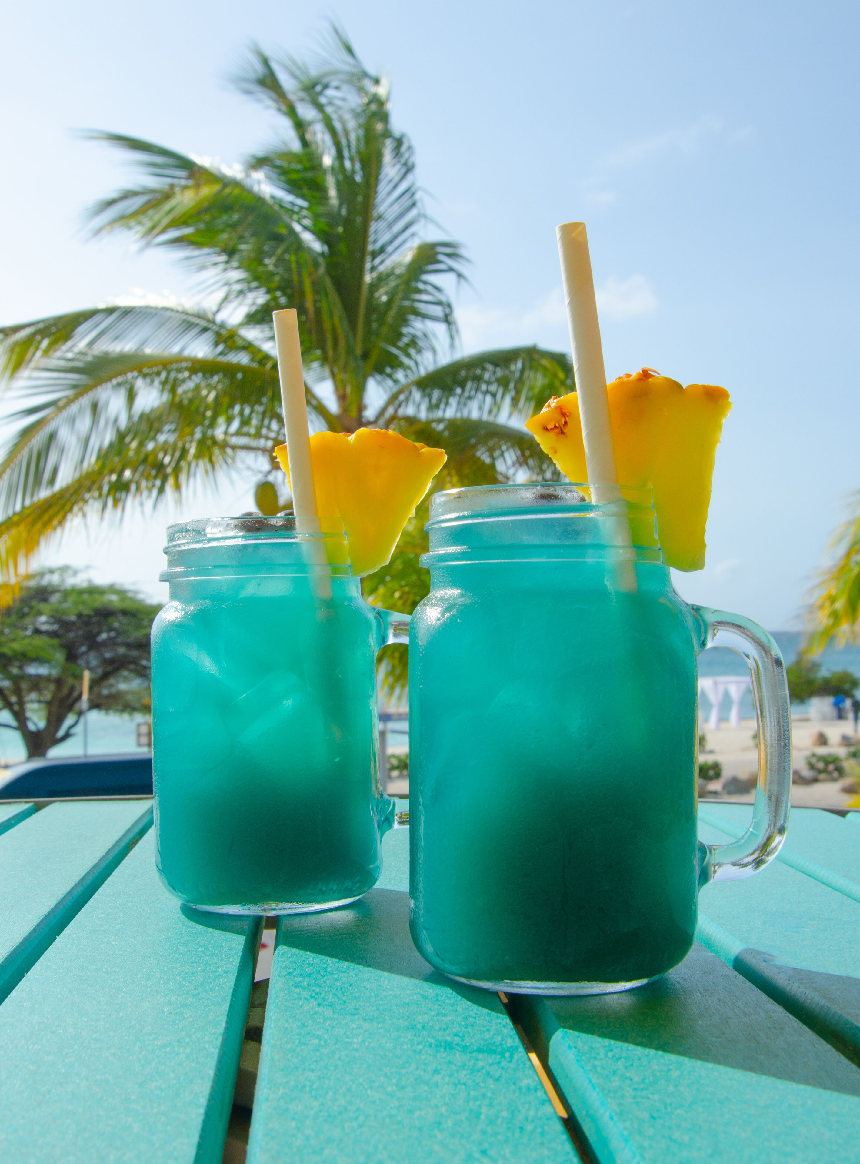 Dos bebidas turquesas con pajitas y trozos de piña sobre una mesa en la playa.