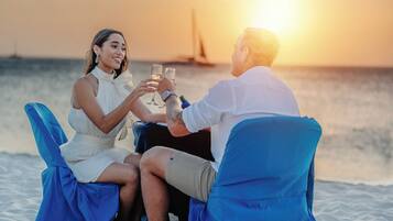 Una pareja sentada en una silla de playa mirando al océano, con un atardecer de fondo.