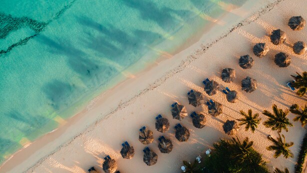 An aerial view of a beach with clear turquoise waters, a sandy shore, and rows of thatched umbrellas and lounge chairs. - Amsterdam Manor Beach Resort (Noord)