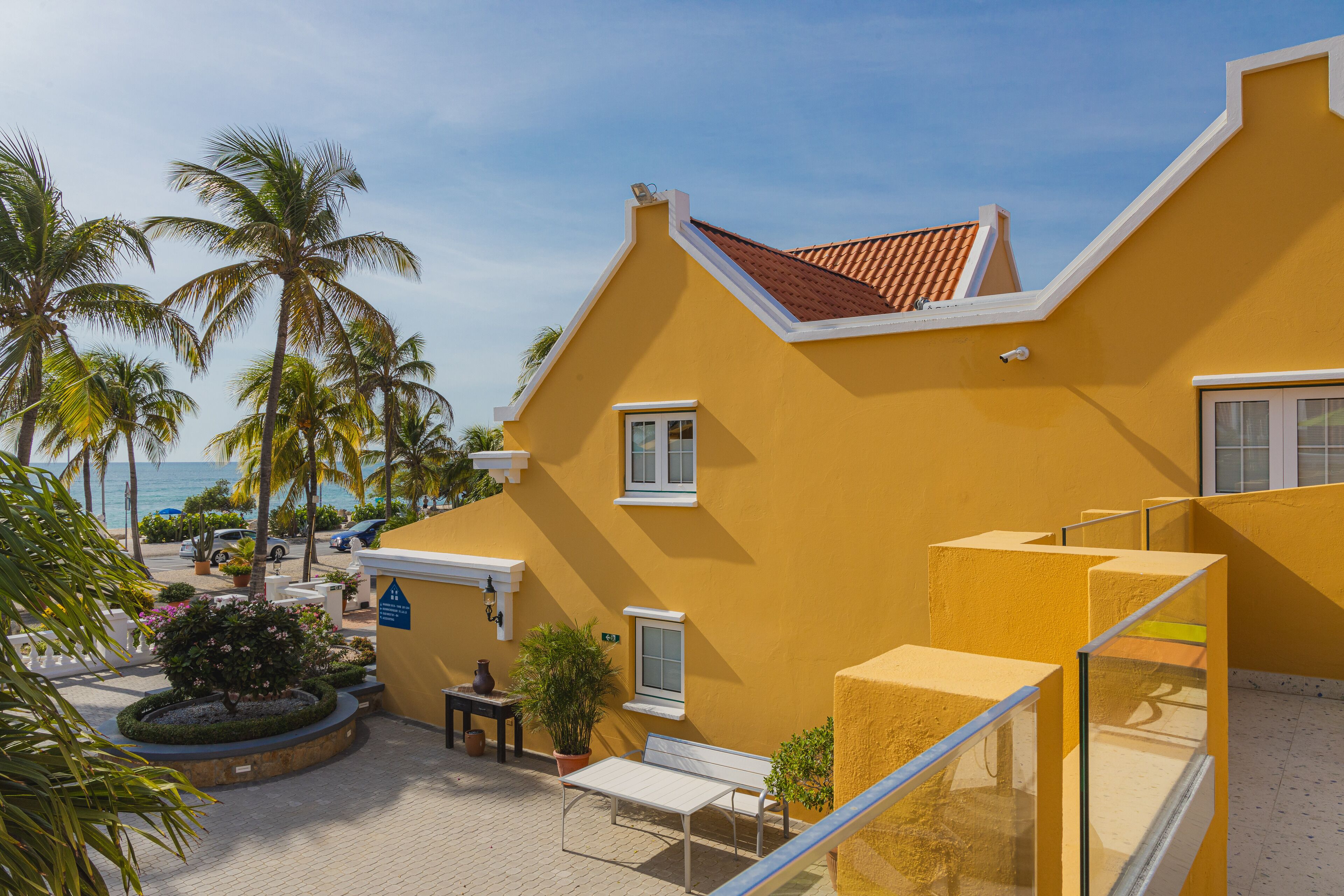 A yellow building with a sloped roof, a small patio with a table and chairs, and palm trees in the background.