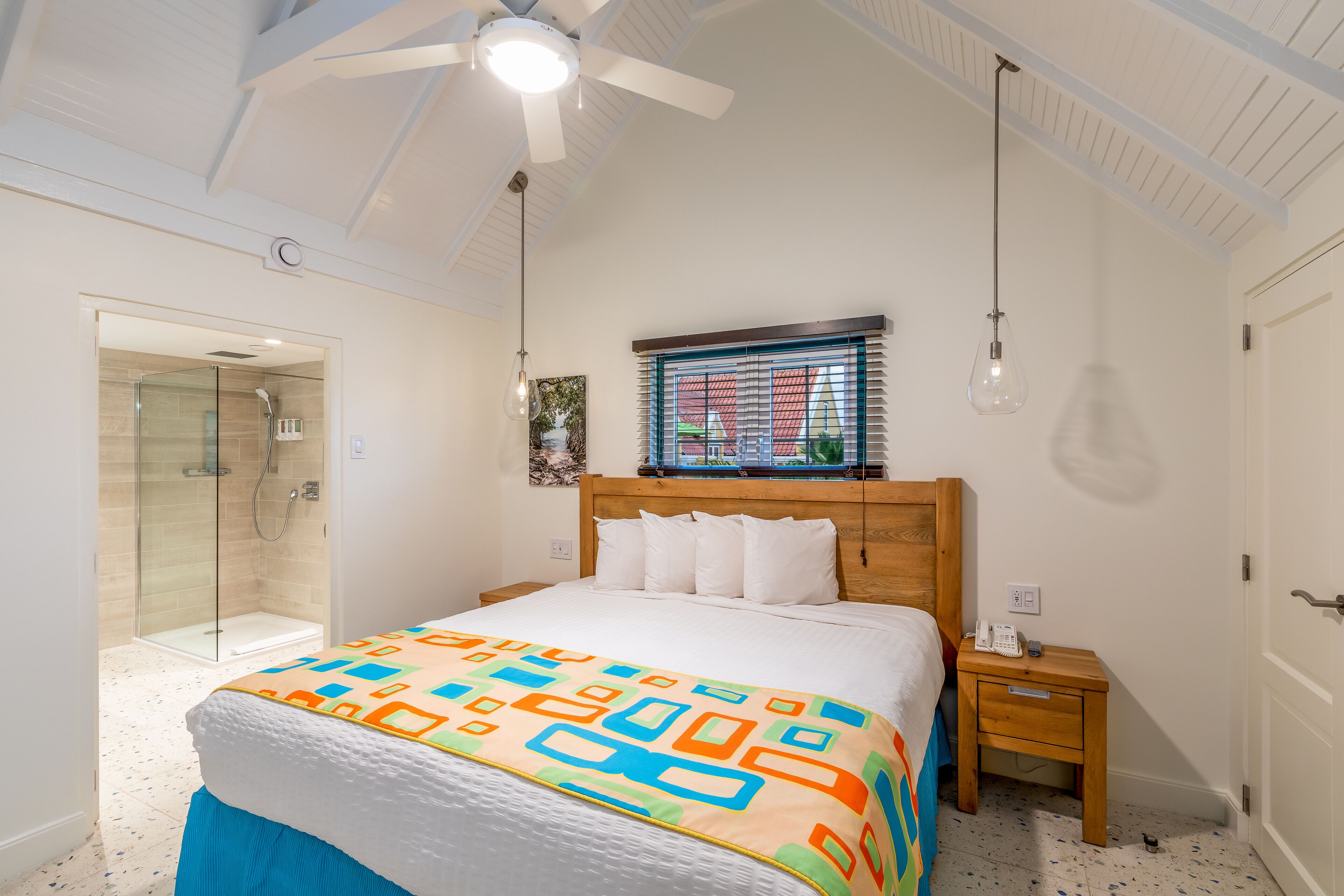 Un dormitorio con cama, mesita de noche, ventilador de techo, ventana con persianas y zona de ducha.