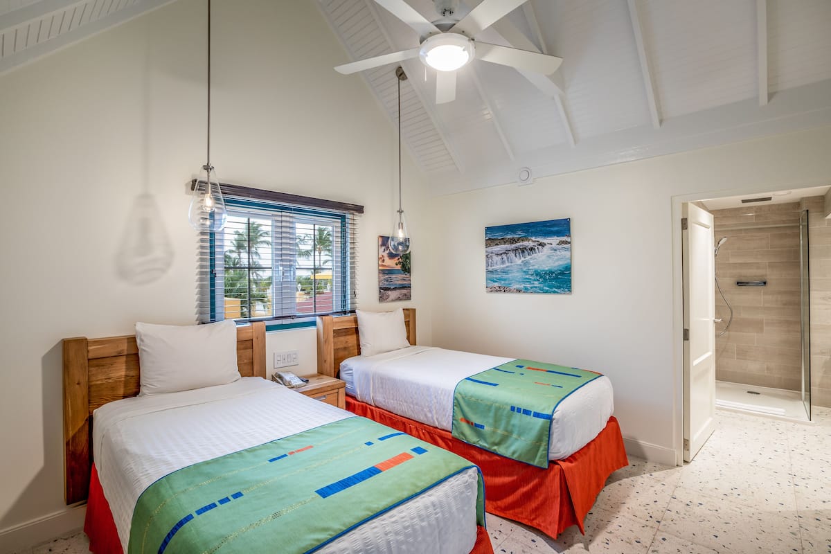 A hotel room with two beds, a shower, a window with blinds, and a ceiling fan.