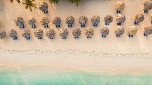 An aerial view of a beach with rows of thatched umbrellas and blue lounge chairs, a sandy shoreline, and turquoise water. - Amsterdam Manor Beach Resort (Noord)