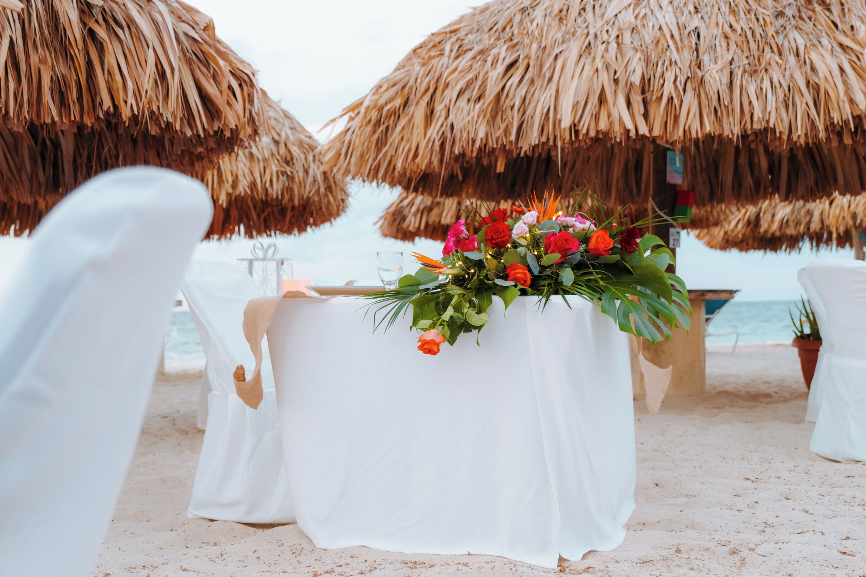 Een trouwdecoratie op het strand met een witte tafelkleed, een bloemstuk als middelpunt en rieten parasols.