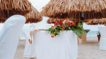 Een trouwdecoratie op het strand met een witte tafelkleed, een bloemstuk als middelpunt en rieten parasols.