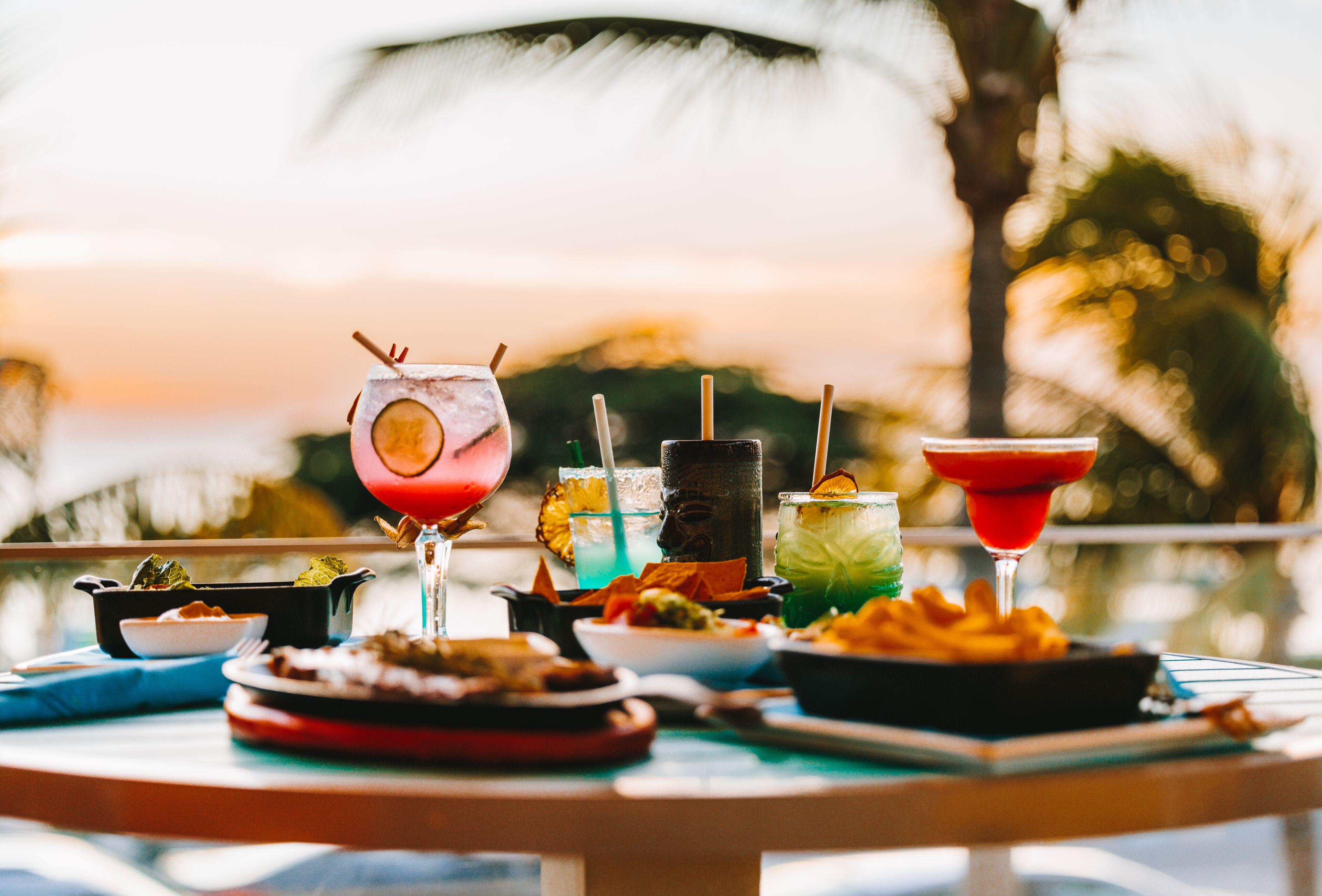 A table with various dishes and drinks, likely a buffet or meal setting, with a view of a beach and palm trees in the background.
