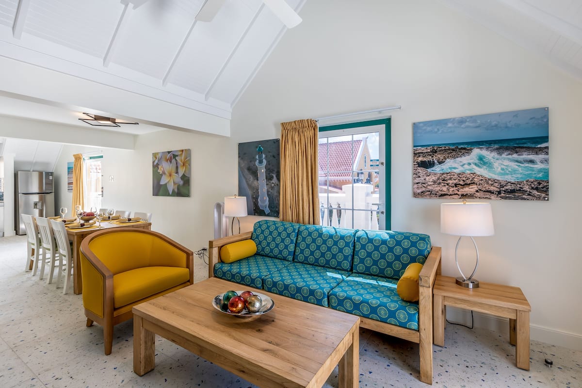 A living room with a blue patterned sofa, a yellow armchair, a wooden coffee table with a bowl of fruit, a lamp, and a painting of a rocky coastline.