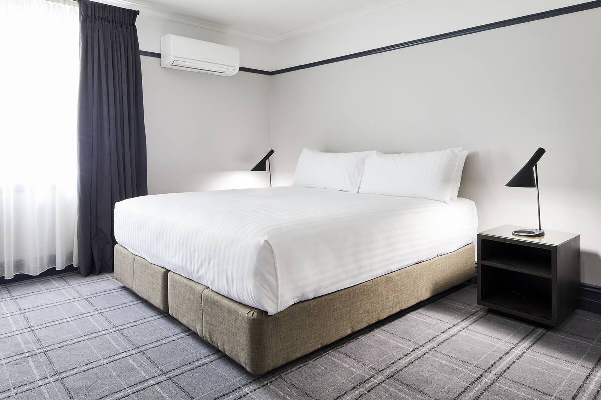 A neatly made bed with white linens, a dark-colored nightstand with a lamp, and a plaid-patterned carpet.