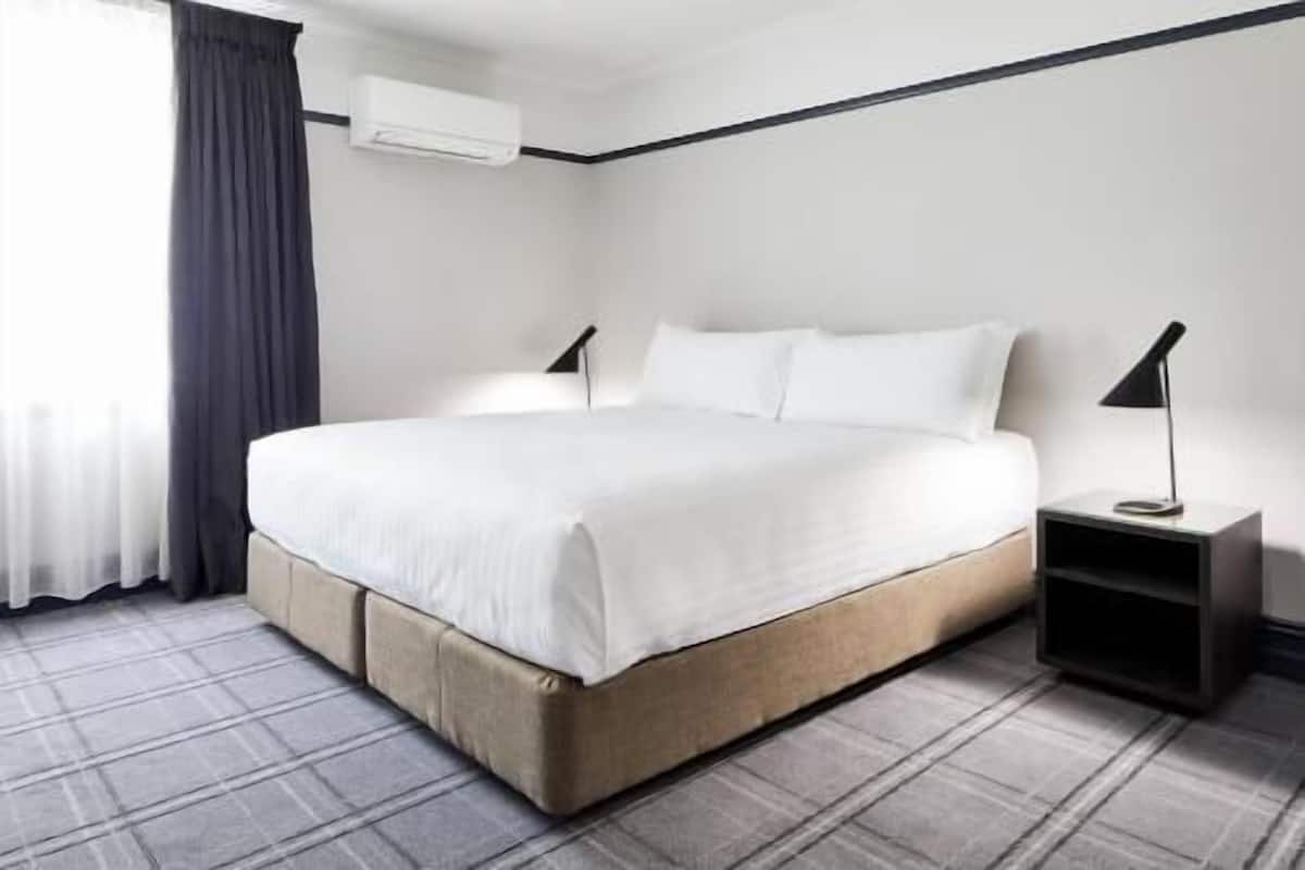 A hotel room with a bed, bedside tables, a nightstand, a window with curtains, and an air conditioning unit.