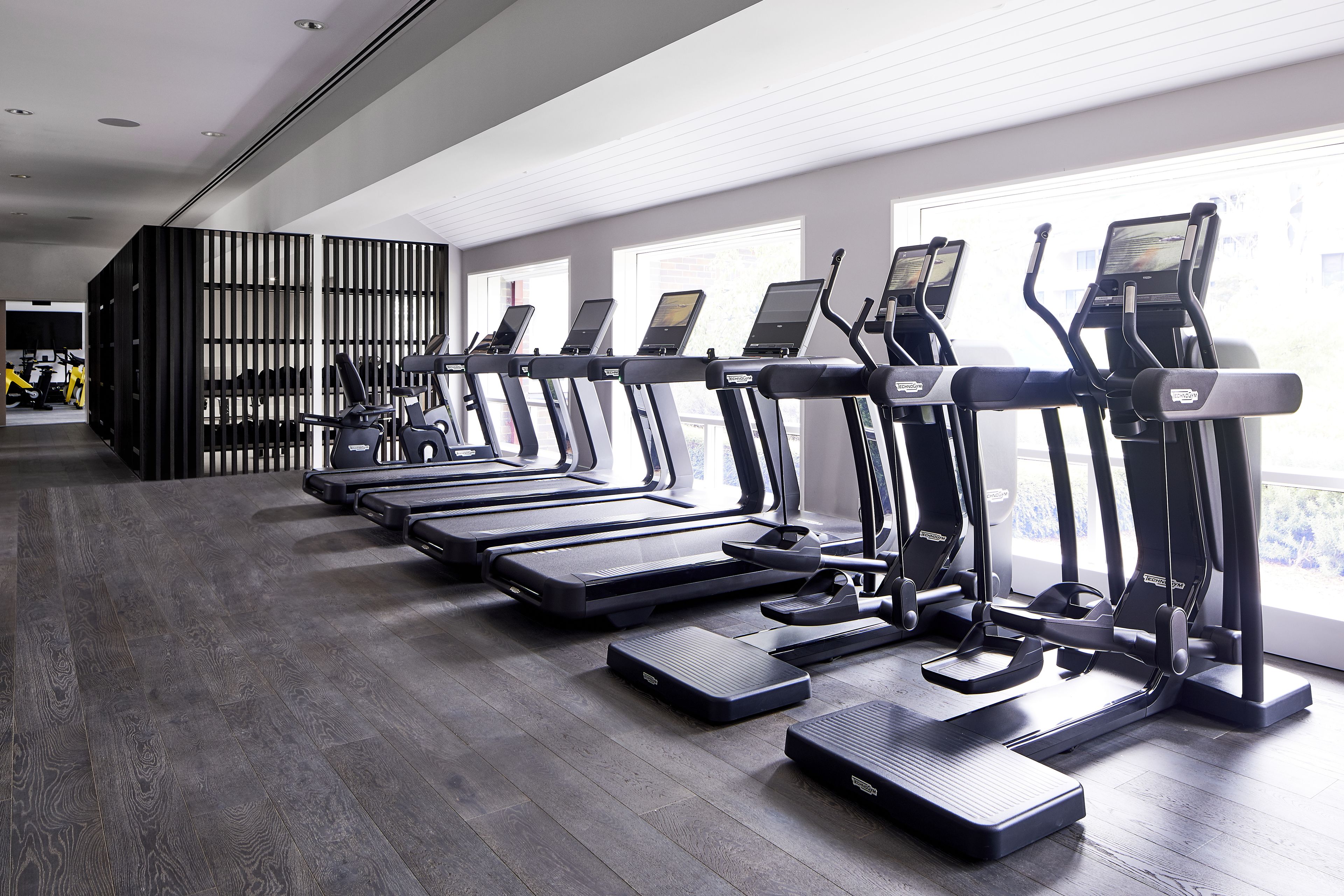 A row of treadmills in a gym with a modern design, large windows, and a dark wooden floor.