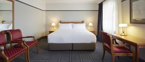 A hotel room with a large bed, two bedside tables with lamps, a desk with a chair, and a large mirror.