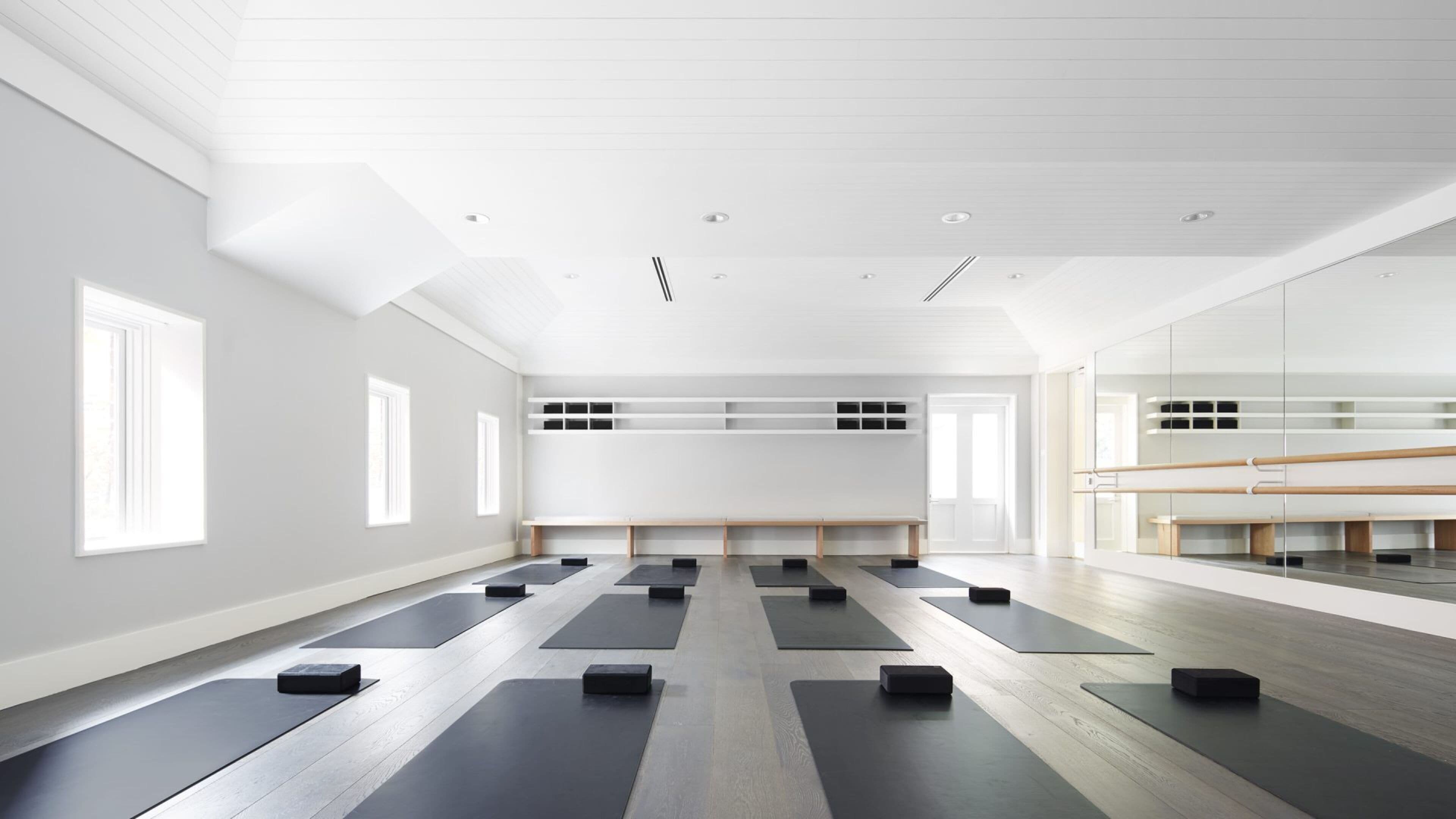 A spacious room with multiple yoga mats arranged in rows, a large mirror on one wall, and a wooden floor.