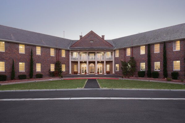 A large brick building with a symmetrical facade, a central entrance with a balcony, and a well-maintained lawn.