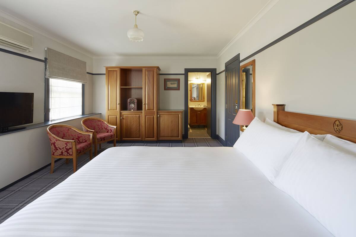 A hotel room with a large bed, a TV, a wardrobe, a desk with a chair, and a bathroom visible through an open door.