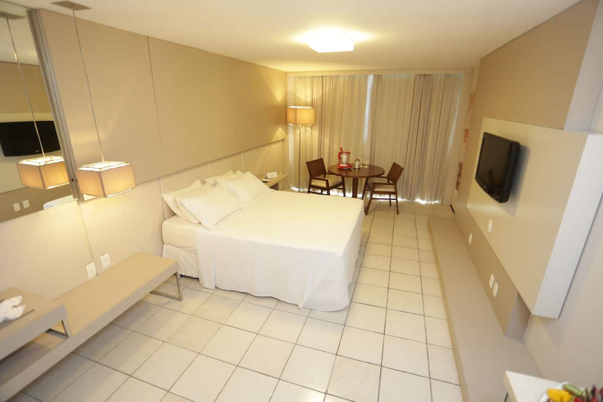 A hotel room with a large bed, a bedside table, a desk with a chair, a TV, and a window with curtains.