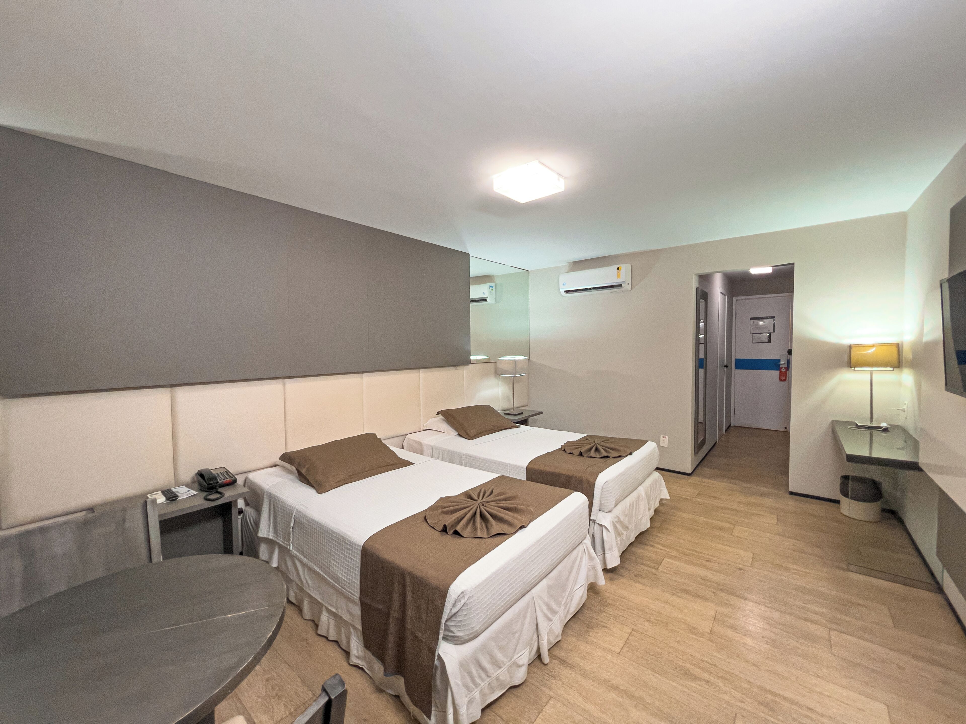 Quarto de hotel com duas camas, uma escrivaninha, uma cadeira, um abajur e um espelho.