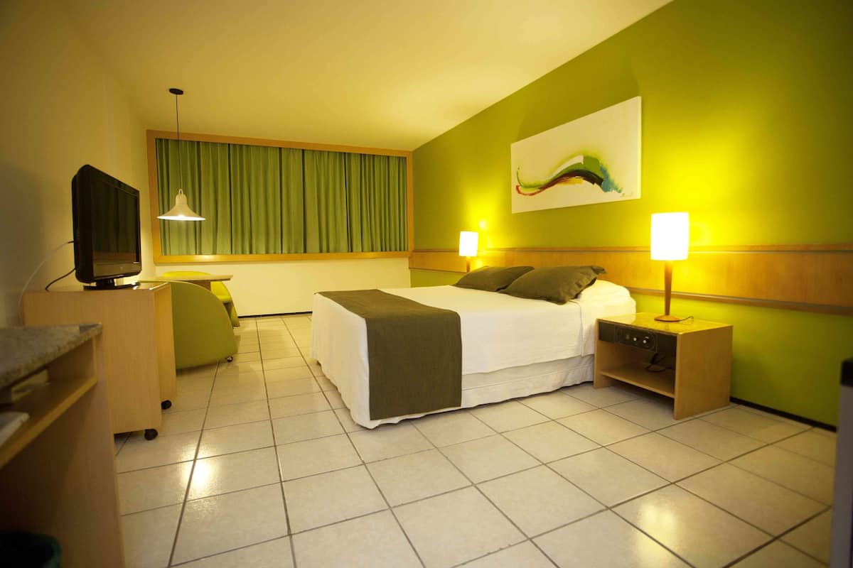 A hotel room with a bed, bedside tables, a desk, a television, and a window with curtains.