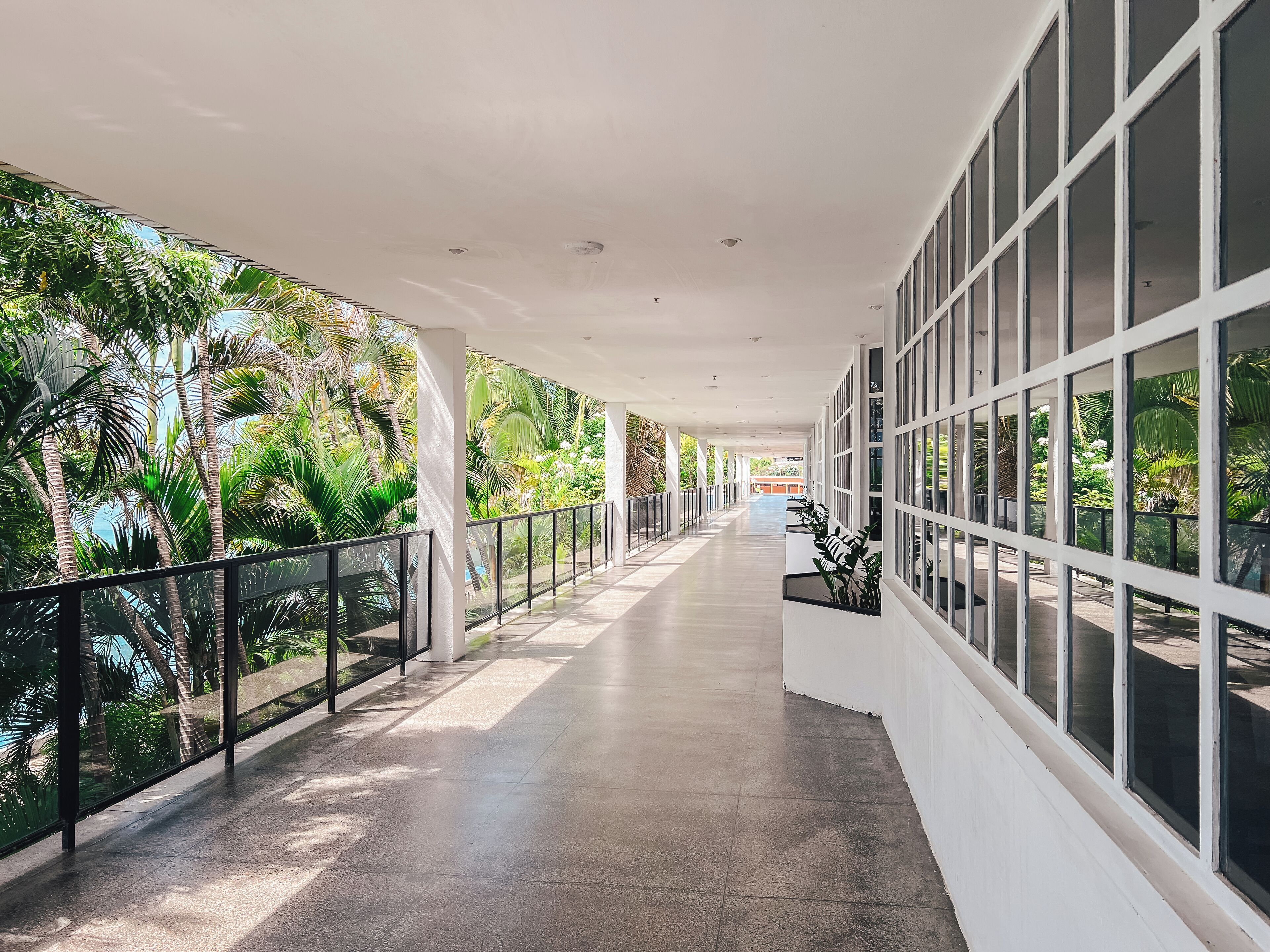 Um corredor coberto com piso ladrilhado, grandes janelas e vista para a vegetação tropical.
